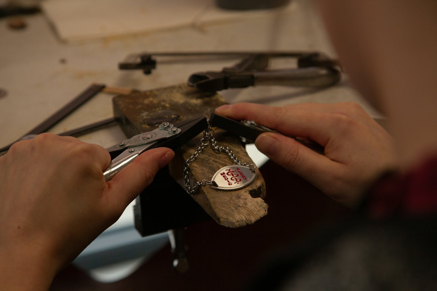 Hands using pliers to craft a delicate metal necklace on a workbench.